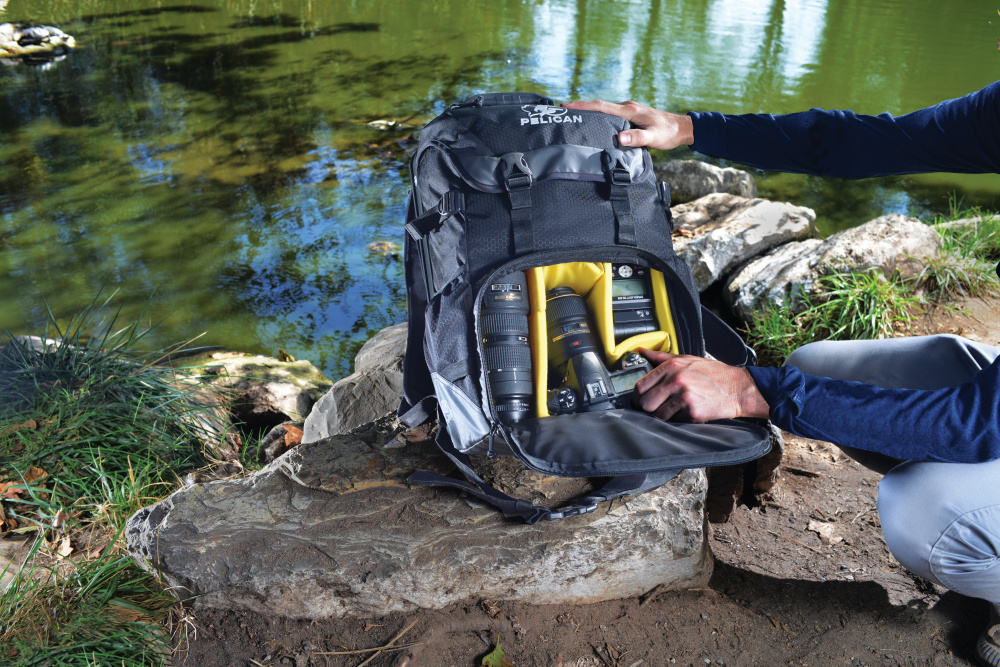 Person at a lake opening a protective backpack to display camera equipment inside.