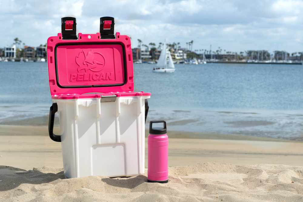 White and pink colored rugged cooler on a beach, next to a bright pink drinkware tumbler.